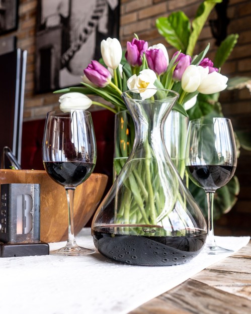 A table setting with a glass wine decanter, two wine glasses, and a vase of tulips in a cozy, stylish environment.