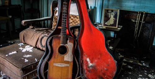 An old acoustic guitar and its red case sit on a worn chair, surrounded by scattered debris and a framed photo in the background.