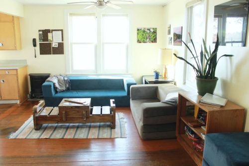 Cozy living room with a blue couch, gray armchair, wooden coffee table, and plants, featuring large windows and warm lighting.