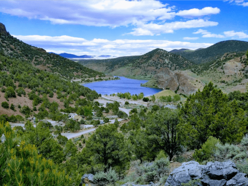 A scenic view of a lake surrounded by green hills and trees under a blue sky with scattered clouds.