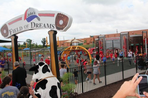 A playground named "Jake's Field of Dreams" with children playing and adults watching, featuring colorful equipment and a cow mascot.