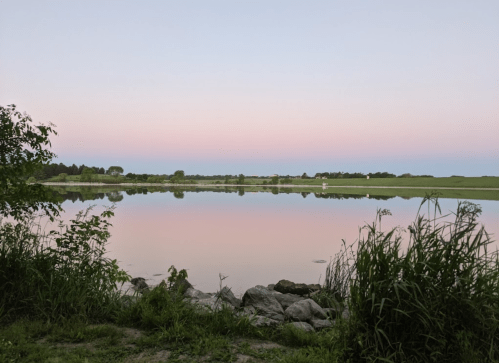 A serene lake at dusk, reflecting soft pastel colors in the sky, surrounded by lush greenery and rocks.