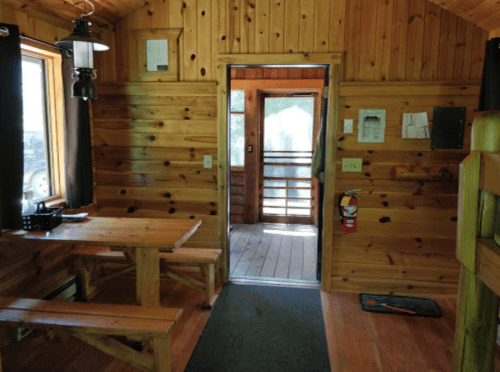 Interior of a wooden cabin with a table, benches, and an open door leading to a porch. Natural light filters in.