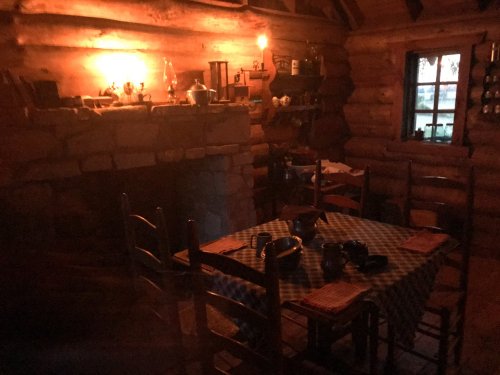 Cozy log cabin interior with a wooden table, chairs, and a warm glow from a lantern on the wall.