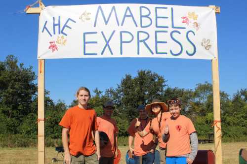 A group of five people in orange shirts stand under a sign that reads "The Mabel Express" in a sunny outdoor setting.