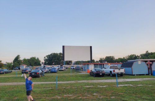 A drive-in movie theater with cars parked and a large blank screen, set against a clear sky. A child is walking nearby.
