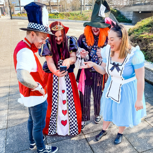 A group of four people in colorful costumes, including a queen, a mad hatter, and Alice, looking at a phone together.