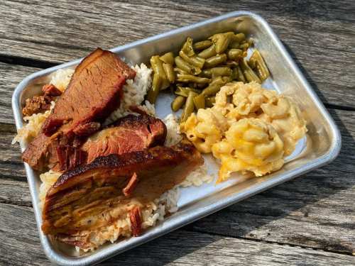 A metal tray with sliced smoked meat, rice, green beans, and macaroni and cheese on a wooden surface.