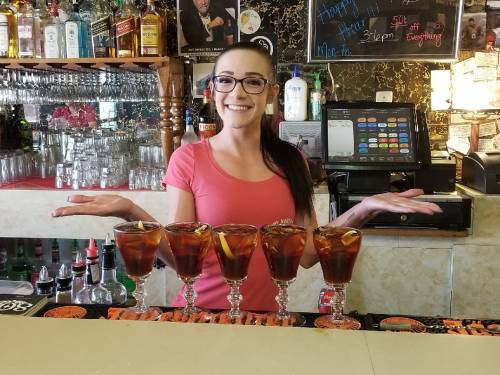 A smiling bartender stands behind a bar, showcasing five drinks with lemon slices in elegant glasses.