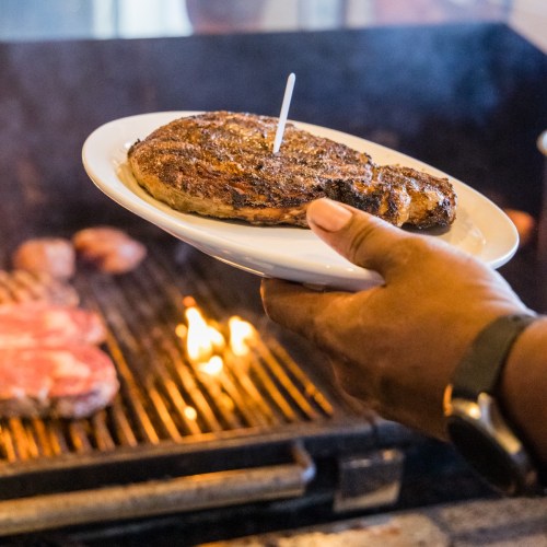A hand holds a plate with a grilled steak, with flames and other meats visible on the grill in the background.