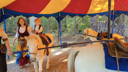 A woman in a costume guides a young boy riding a pony under a colorful tent in a forested area.