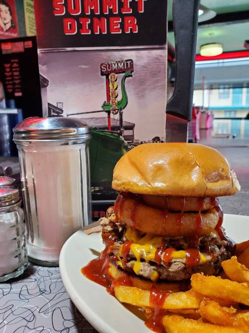 A towering burger with cheese, onion rings, and ketchup, served with fries, next to a diner menu and condiments.