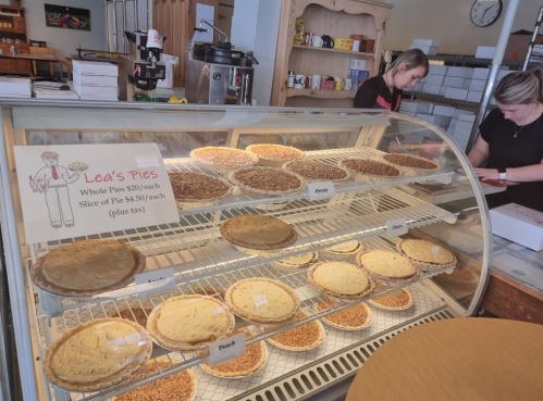 Display case with various whole pies and slices at Lea's Pies, including pecan and cream pies, with staff in the background.