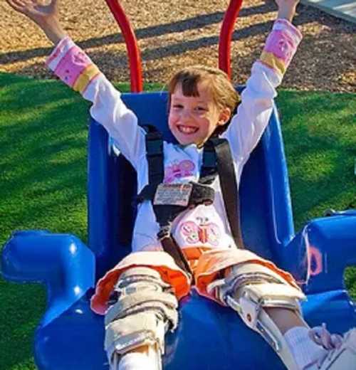 A smiling child in a swing, wearing a colorful outfit and safety harness, joyfully raising their arms in excitement.