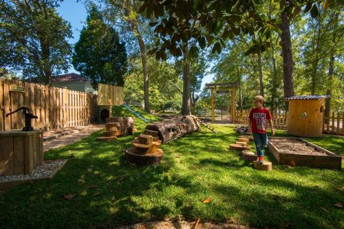 A child explores a nature-themed playground with logs, a sandbox, and a wooden playhouse surrounded by trees.