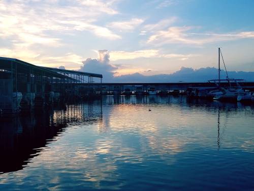 A serene marina at sunset, with boats reflected in calm water and colorful clouds in the sky.