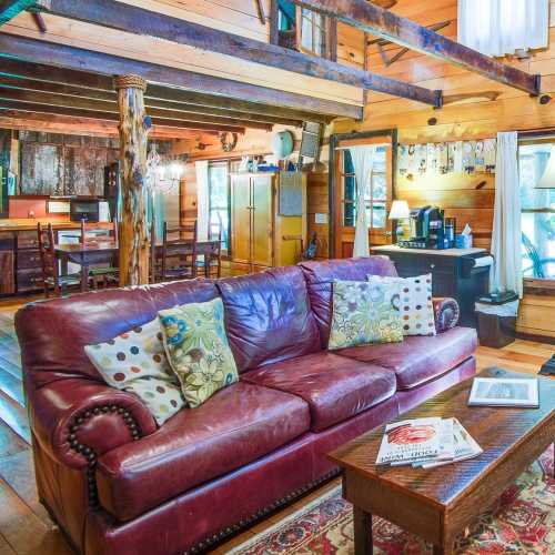 Cozy living room with a burgundy leather sofa, decorative pillows, wooden beams, and a rustic dining area in the background.