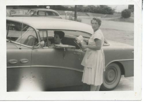 A woman stands by a vintage car, handing something to a person inside, in a black-and-white photo.