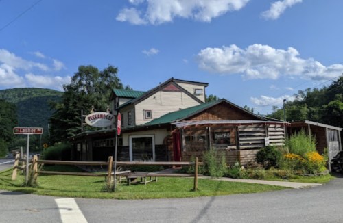 A rustic building with a green roof, surrounded by greenery and flowers, under a blue sky with scattered clouds.