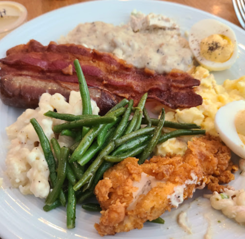 A plate of breakfast featuring bacon, sausage, scrambled eggs, green beans, fried chicken, and a sliced hard-boiled egg.