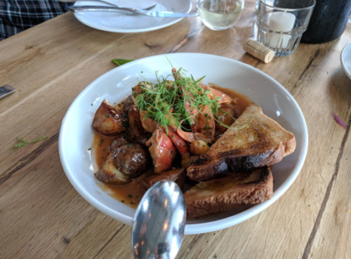 A bowl of sautéed vegetables and seafood, garnished with herbs, served with toasted bread on a wooden table.