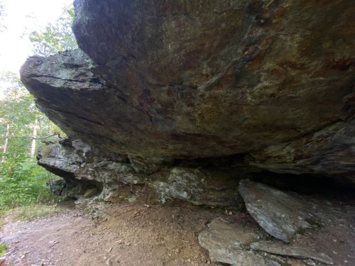 A large rock overhang in a wooded area, with a dirt path leading underneath it.