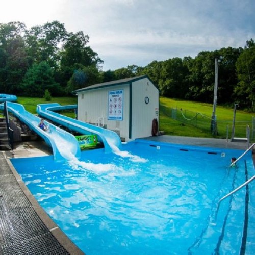 A water park scene featuring two slides leading into a pool, surrounded by greenery and a small building nearby.