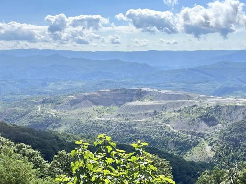 A panoramic view of a mountainous landscape with a quarry, lush greenery, and a cloudy blue sky.