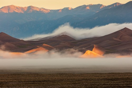 A serene landscape of sand dunes bathed in golden light, with mountains and mist in the background.
