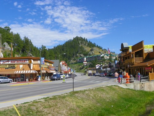 A scenic view of a small town street lined with shops and restaurants, surrounded by green hills and blue skies.