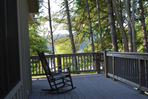 A wooden deck with a rocking chair, surrounded by tall trees and a view of a serene landscape in the background.