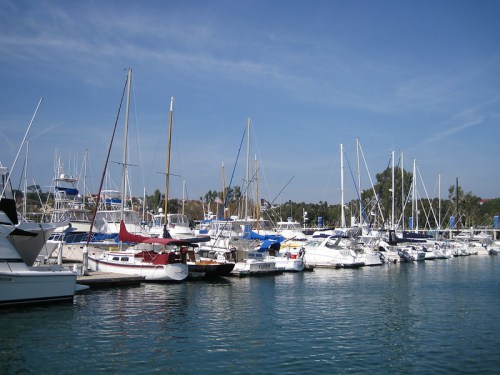 A marina filled with various sailboats and yachts, surrounded by clear blue water and a bright sky.