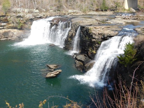 A scenic view of two waterfalls cascading into a clear blue pool, surrounded by rocky terrain and sparse trees.