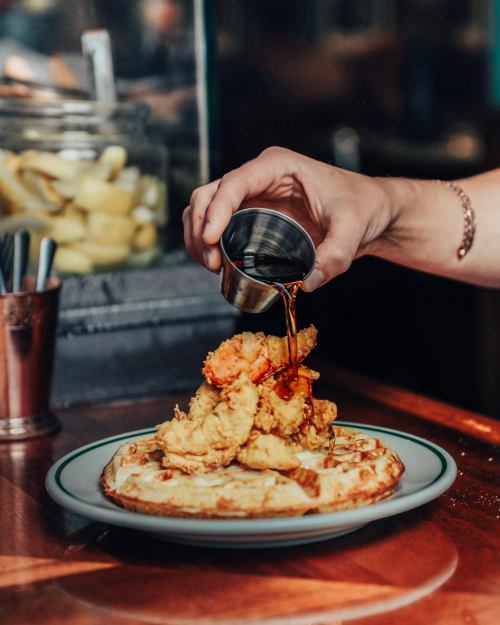 A hand pours syrup over a plate of fried shrimp on a waffle, set on a wooden table.