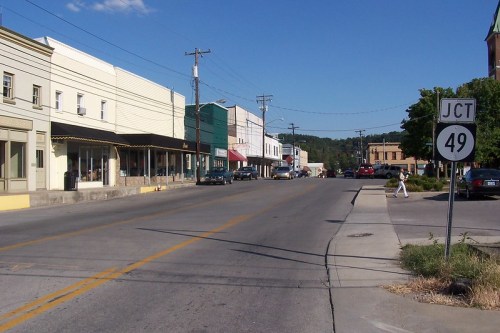 A quiet street scene with storefronts, parked cars, and a road sign indicating Junction 49 under a clear blue sky.
