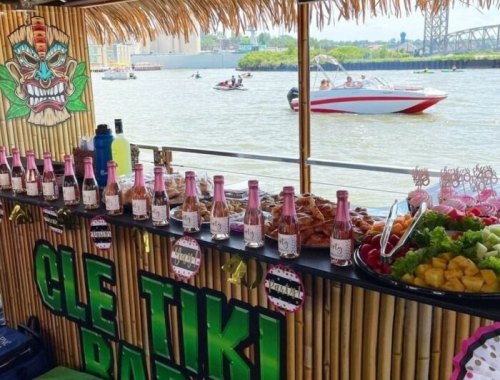 A tiki bar setup with drinks and snacks, overlooking a river with boats in the background. Tropical decor and vibrant colors.