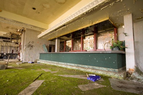 Abandoned interior with a moss-covered reception desk, peeling walls, and scattered debris in a dilapidated space.