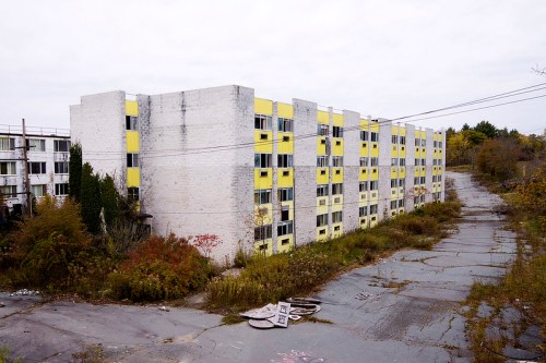 Abandoned building with yellow accents, surrounded by overgrown vegetation and a cracked asphalt road.
