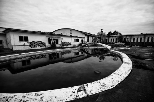 Abandoned building with graffiti, reflecting in a still, empty pool under a cloudy sky. Black and white photography.