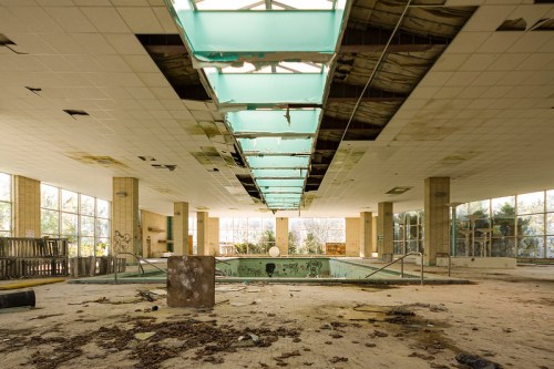 Abandoned indoor pool area with broken skylights, debris, and overgrown vegetation, showcasing decay and neglect.
