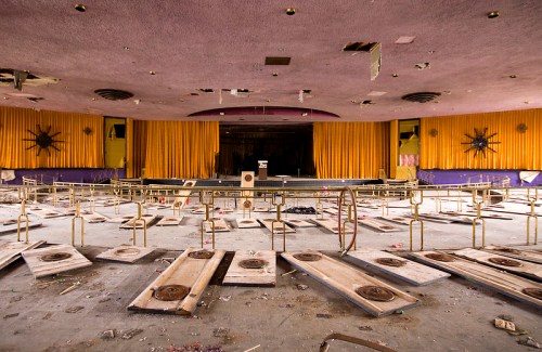 Abandoned theater interior with a stage, yellow curtains, and debris scattered across the floor.