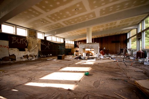 An abandoned room with scattered debris, graffiti on the walls, and large windows letting in natural light.