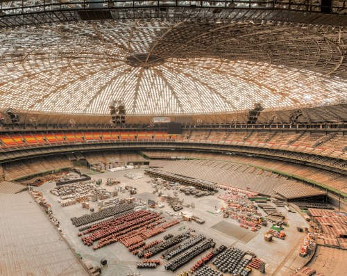 An empty stadium with a large, intricate roof, filled with stacks of materials and equipment on the ground.