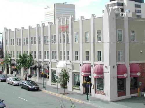 A street view of a multi-story building with shops and restaurants, featuring awnings and greenery along the sidewalk.