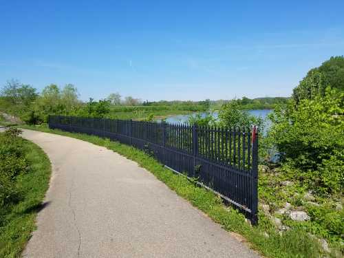 A paved path alongside a black fence, with greenery and a calm lake visible in the background under a clear blue sky.