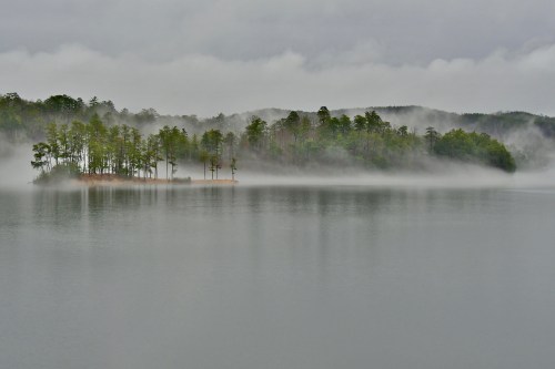 A misty lake scene with an island of trees, surrounded by fog and distant hills under a cloudy sky.