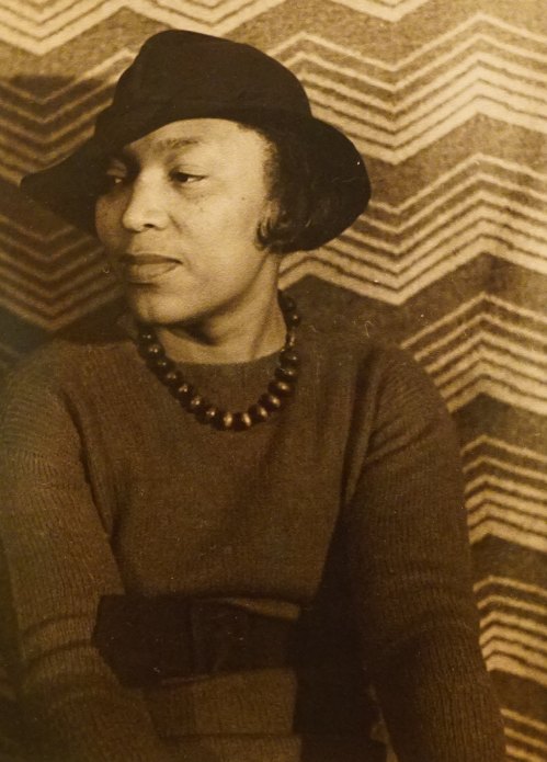A woman in a hat and necklace poses thoughtfully against a patterned background.