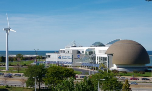 A modern science center with a large dome, solar panels, and a wind turbine, set against a blue sky and lake.