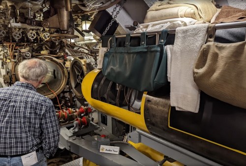 A person examines machinery inside a submarine, with towels and bags hanging nearby.