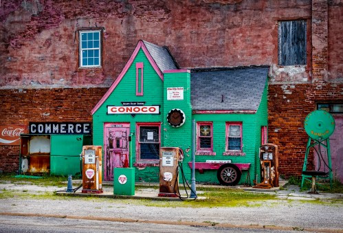 A vintage gas station with a green and pink building, old gas pumps, and a weathered brick wall in the background.